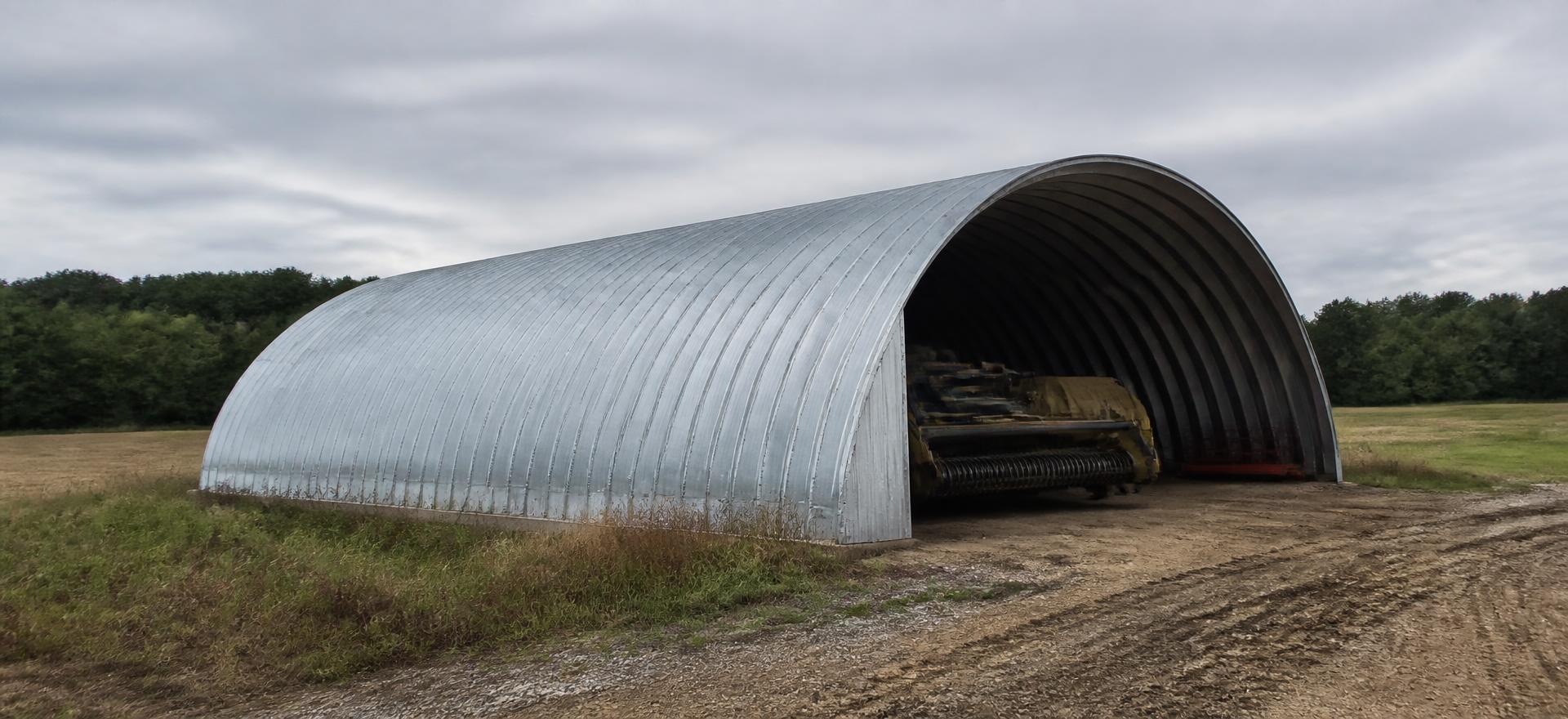 Concept photograph of a galvanized corrugated arch building with an open end sheltering farm equipment beside a cut field; example configuration, not a real project.