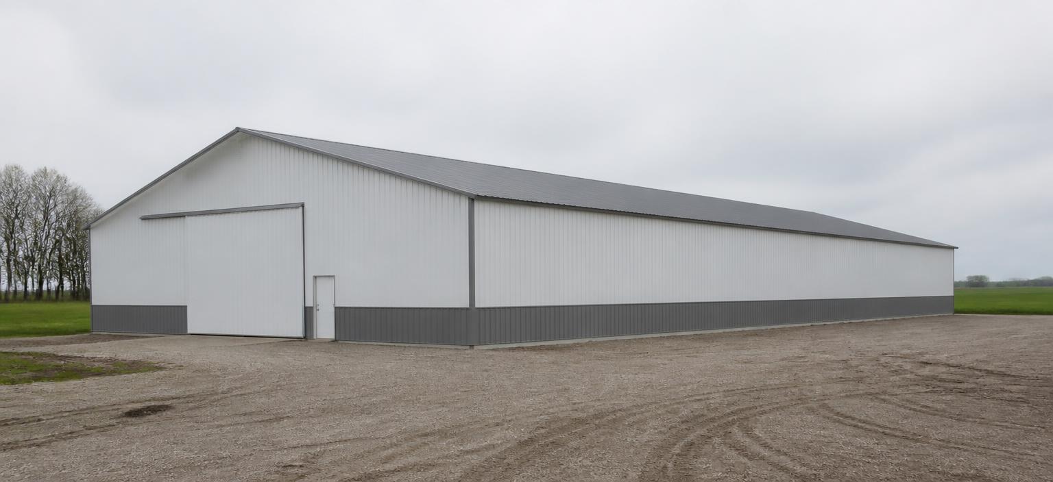 Illustrative steel building configuration showing a wide white agricultural steel building with a grey wainscot, one wide equipment door, one personnel door and a grey ribbed gable roof in a gravel farmyard under overcast sky.