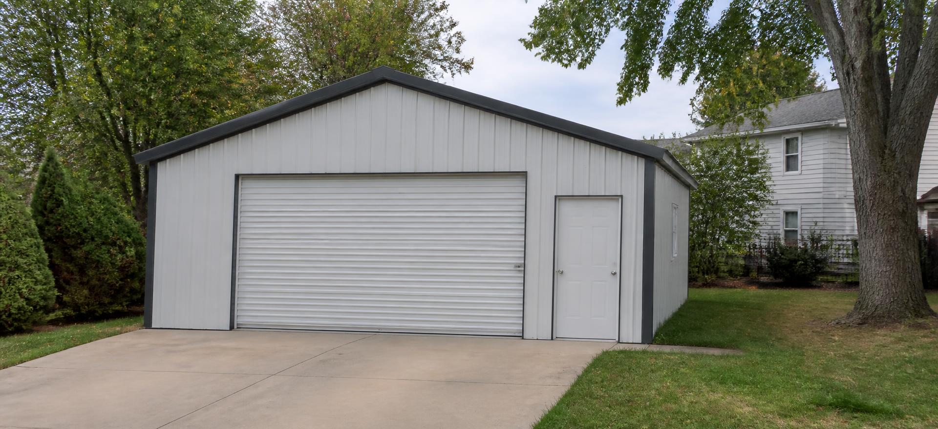 Concept photograph of a small white steel garage with a single wide overhead door and a walk door at the end of a concrete driveway; example configuration, not a real project.