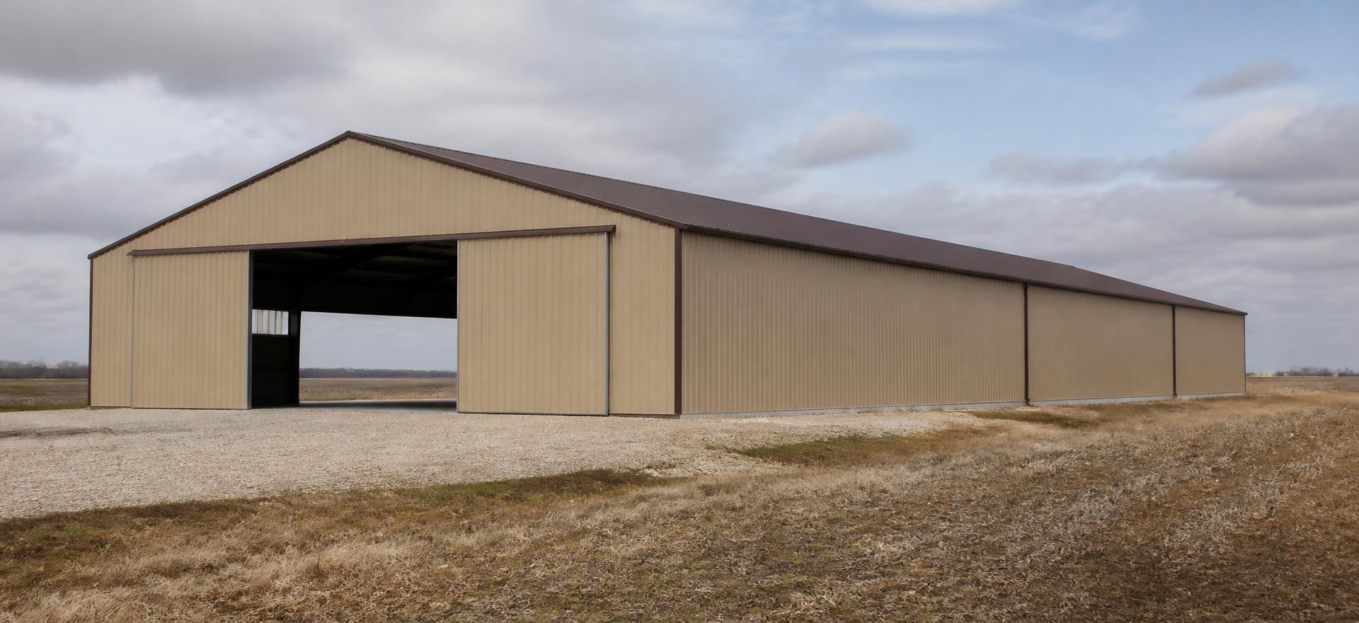 Concept photograph of a long tan rigid-frame agricultural building with a wide open sliding door on the end wall beside a stubble field; example configuration, not a real project.
