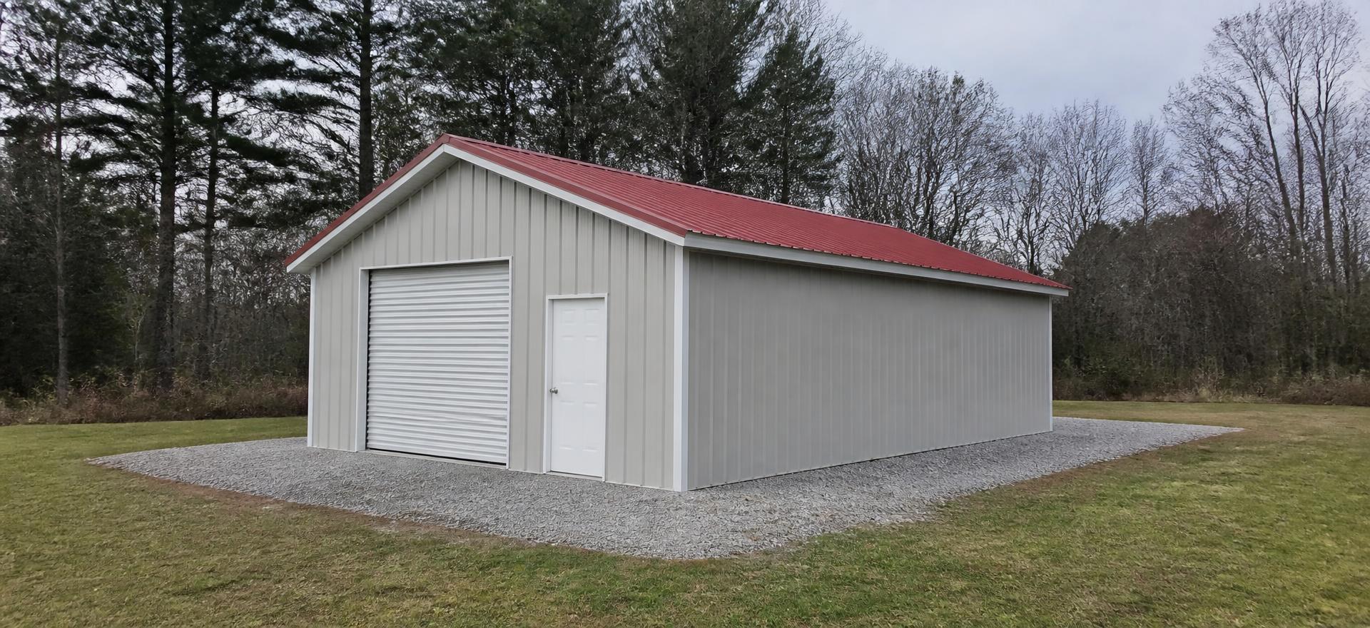 Concept photograph of a 24 by 31 foot enclosed metal garage with a galvanized tube frame, stone-grey vertical-rib walls, a red gable metal roof, one white roll-up door and one walk door on a gravel pad; illustrative configuration, not a real project.