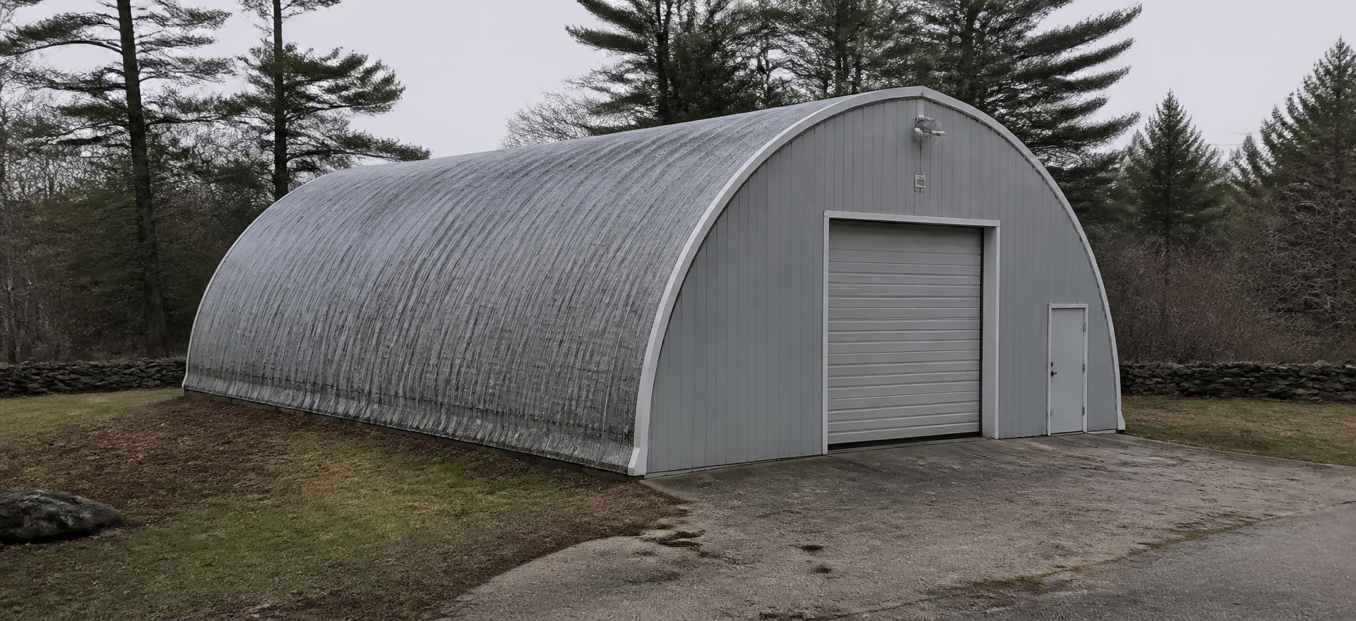 Concept photograph of a galvanized arch steel workshop with a flat grey end wall, one roll-up door and a walk door on a paved apron; example configuration, not a real project.