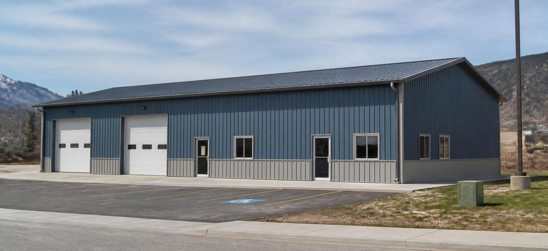 Concept photograph of a 40 by 60 foot blue steel commercial shop with gray wainscot, two overhead doors, glazed entries and a small striped parking area; example configuration, not a real project.
