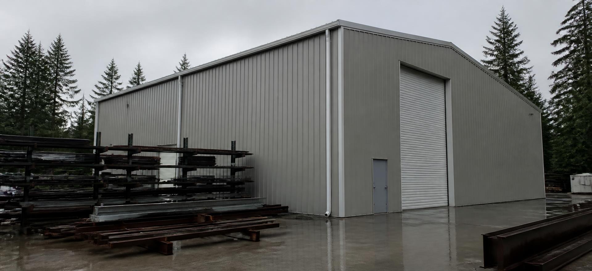 Concept photograph of a tall grey steel industrial building with high sidewalls and one very tall roll-up door beside racked steel stock in the rain; example configuration, not a real project.