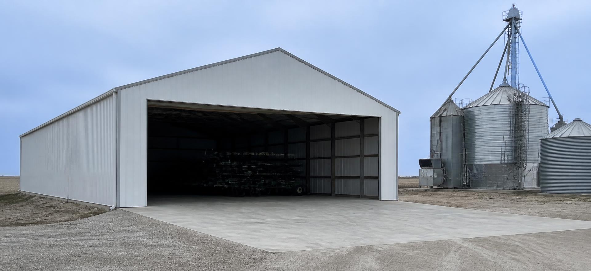Concept photograph of a white clear-span steel farm building with a very wide open end bay on a concrete drive beside grain bins; example configuration, not a real project.