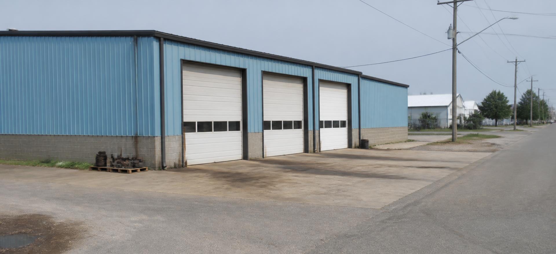 Concept photograph of a light blue steel service building with three roll-up bays over a worn concrete apron on a small industrial street; example configuration, not a real project.