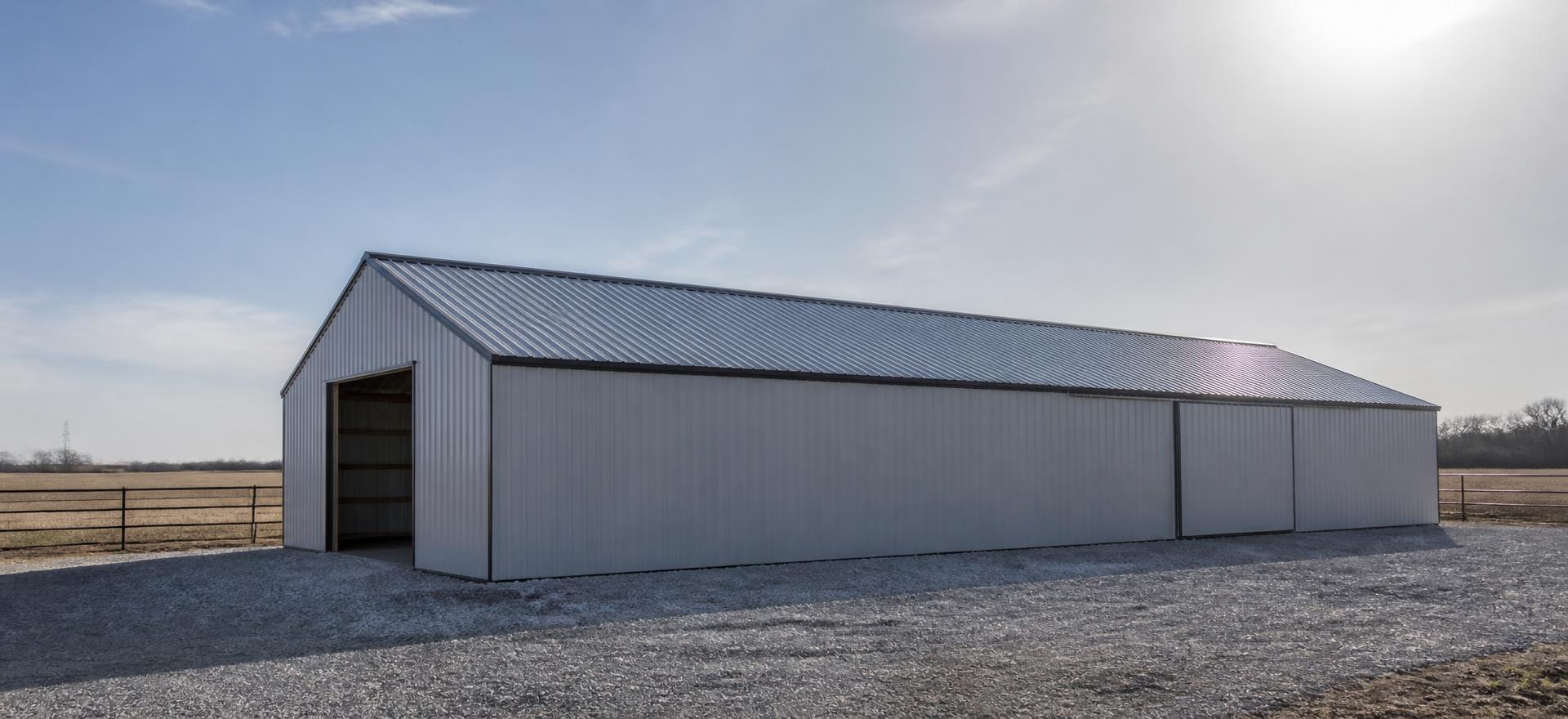 Concept photograph of a silver-grey steel farm building with a long roof plane, one open bay and a sliding door on a fresh gravel pad; example configuration, not a real project.