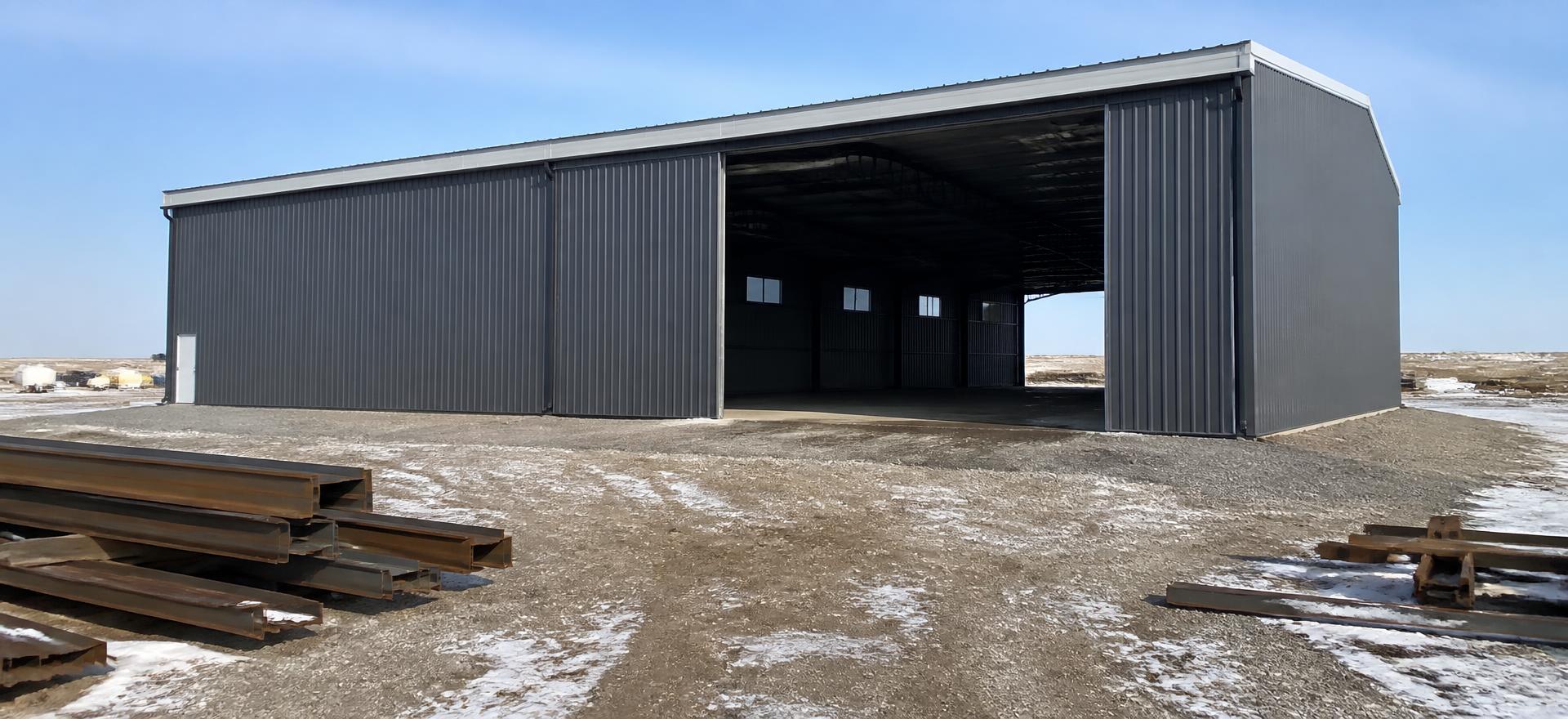 Concept photograph of a dark grey clear-span steel shop with a very wide open sliding door showing an unobstructed interior, on a snow-patched gravel yard; example configuration, not a real project.