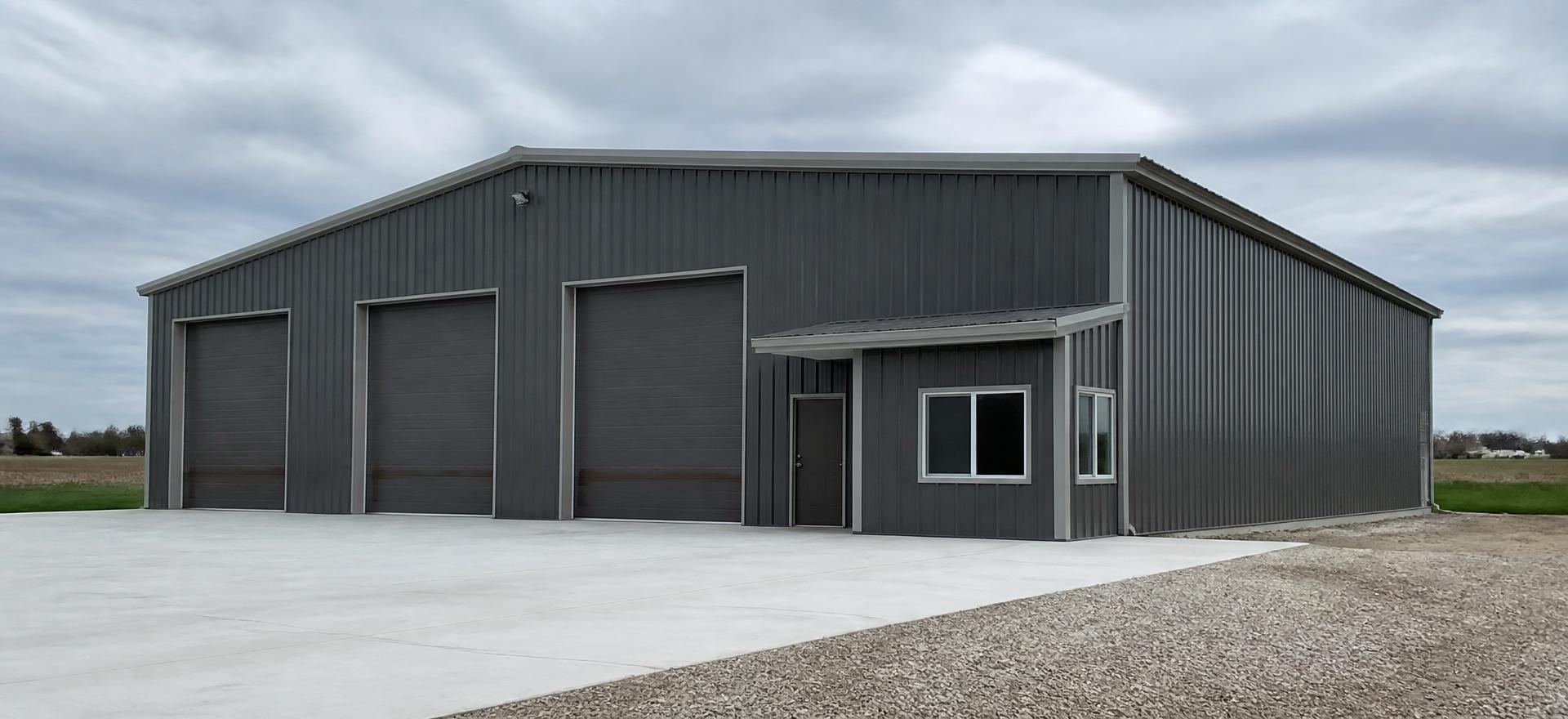 Concept photograph of a charcoal post-frame commercial shop clad in steel with three tall overhead doors and a small window bay on a concrete apron; example configuration, not a real project.