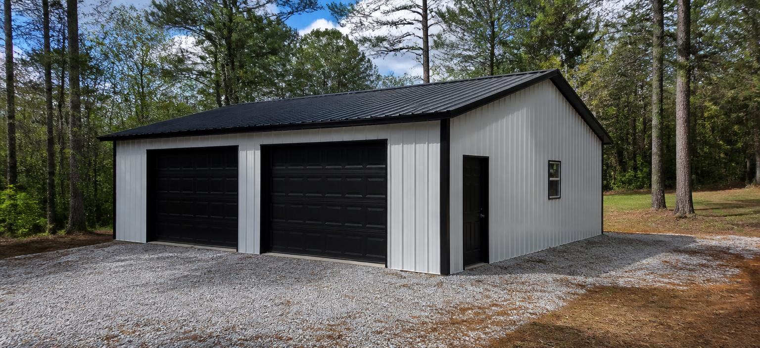 Illustrative steel building configuration showing a small white enclosed metal garage with black trim, two residential-size overhead doors, one personnel door, one small window and a black vertical-rib roof on a gravel pad among pine trees.