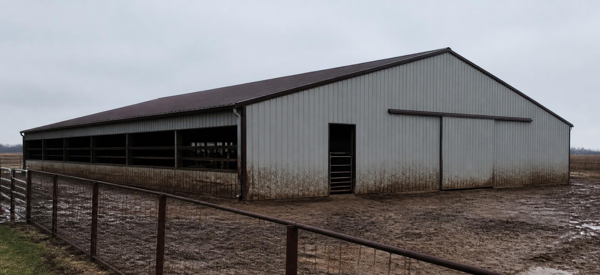 Concept photograph of a grey post-frame livestock barn clad in steel with open ventilated sidewall bays and a wide sliding door beside a muddy lot; example configuration, not a real project.