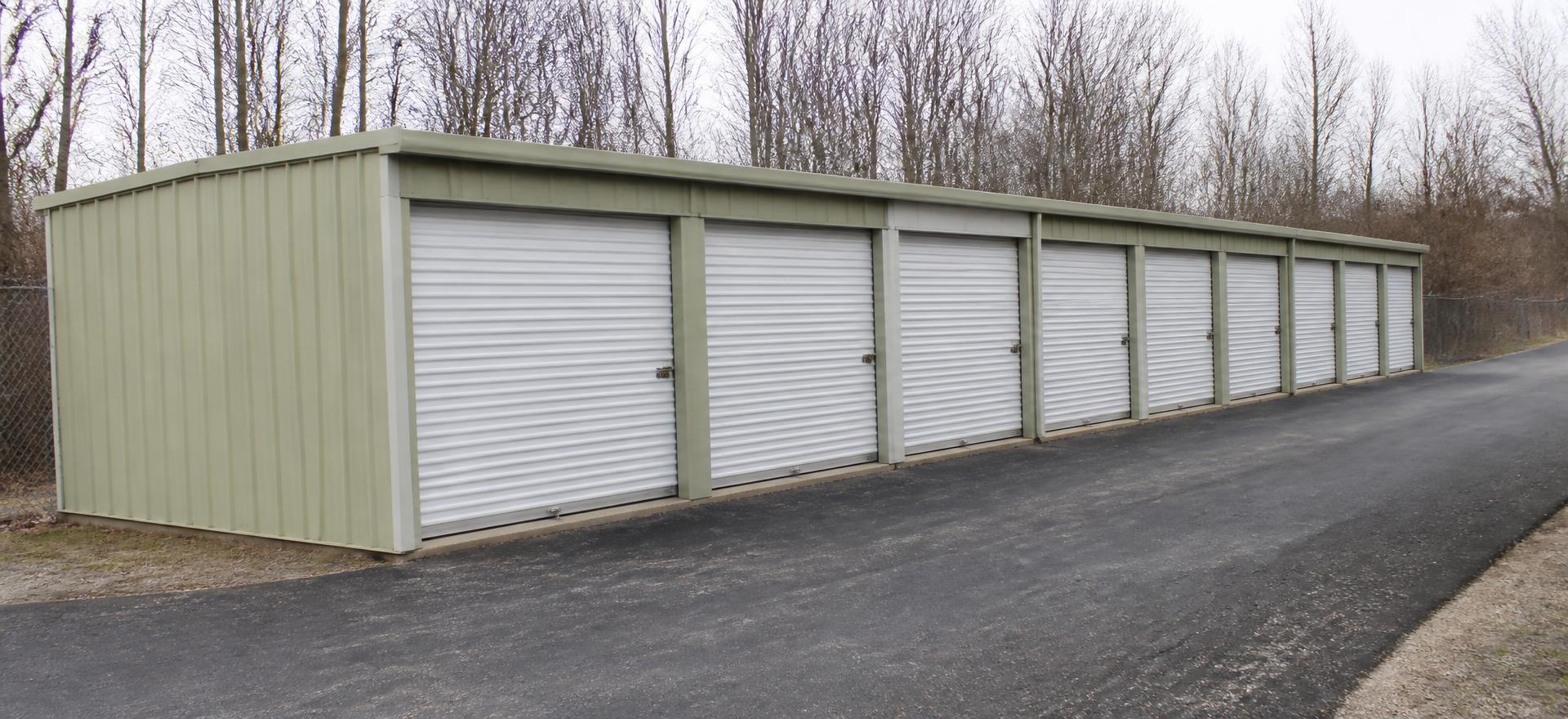 Concept photograph of a single row of pale green steel storage units with white roll-up doors along a narrow asphalt drive; example configuration, not a real project.