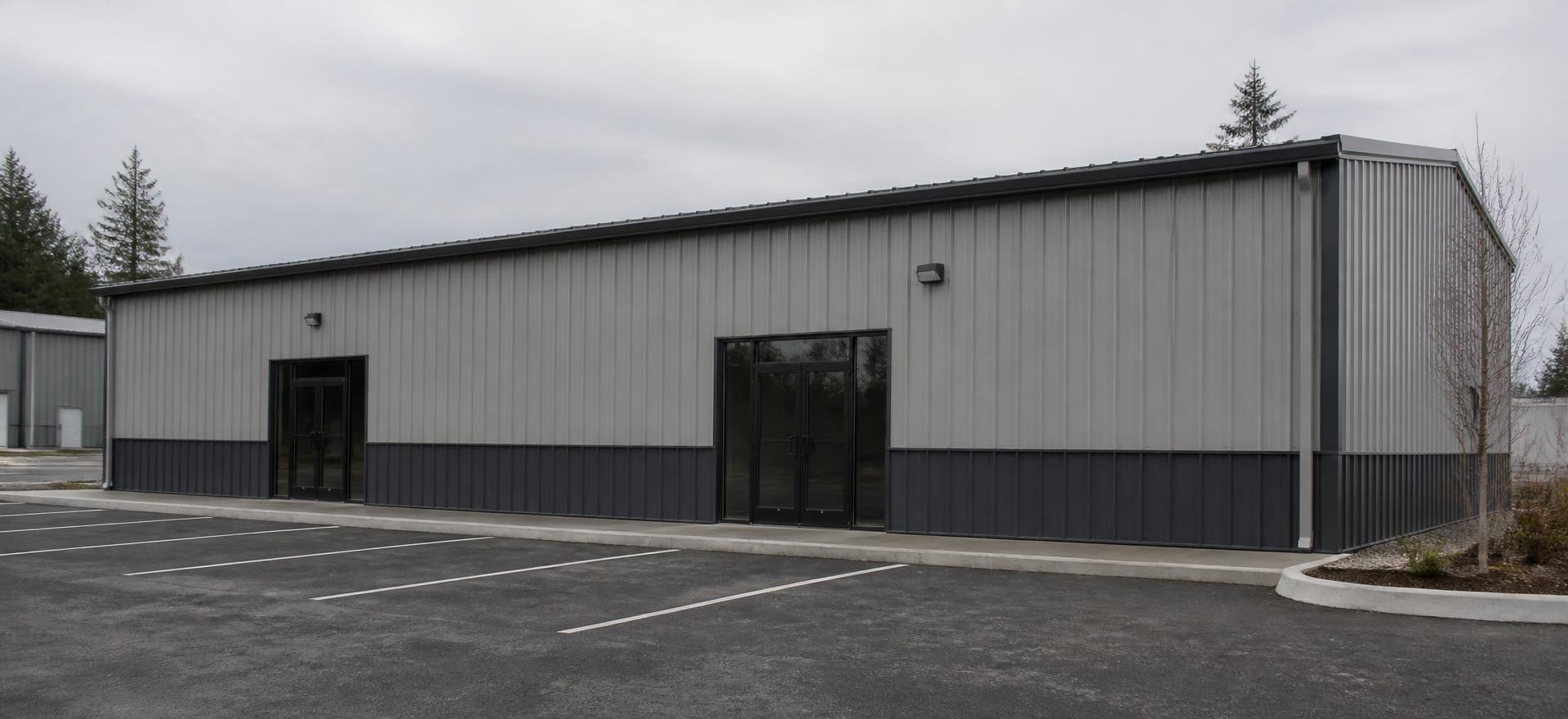 Concept photograph of a grey ribbed steel commercial building with two glass entrances and a dark base wainscot beside a striped parking lot; example configuration, not a real project.