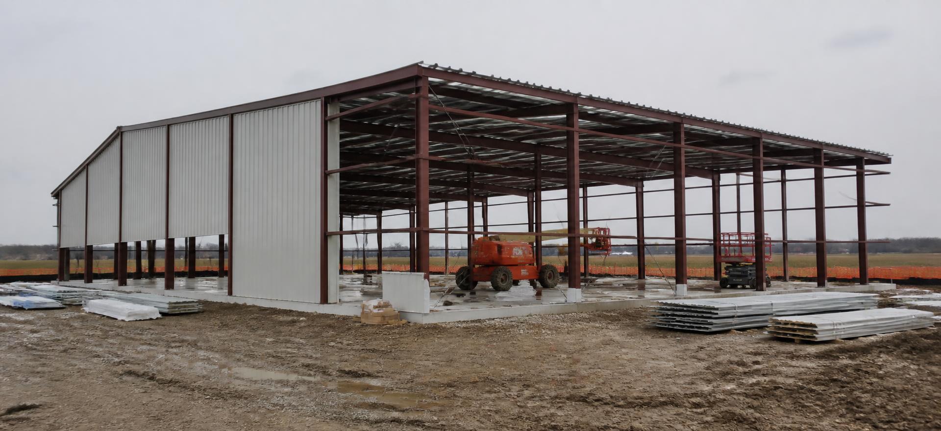 Concept photograph of a 60 by 100 foot steel building kit part-way through erection, with red-oxide primed frames, purlins and half the wall panels installed on a muddy site; example configuration, not a real project.