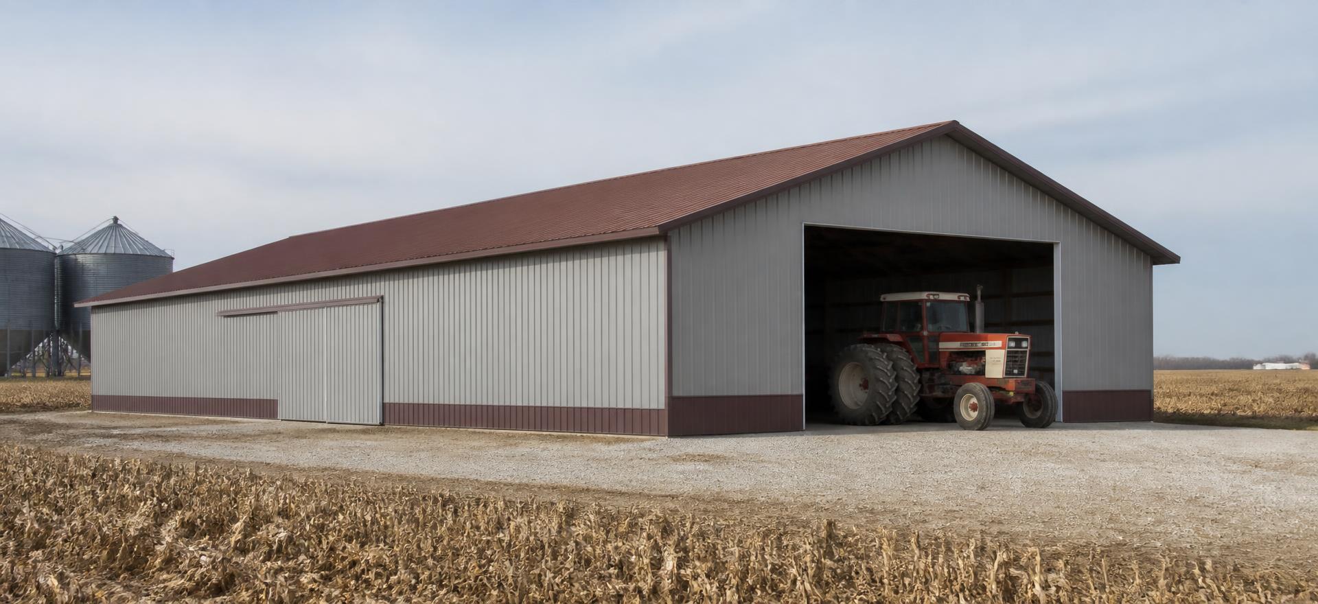 Concept photograph of a 60 by 120 foot grey post-frame machine shed with a red-brown gable metal roof, wide open equipment bay with a tractor inside and a side sliding door beside a harvested field and grain bins; illustrative configuration, not a real project.