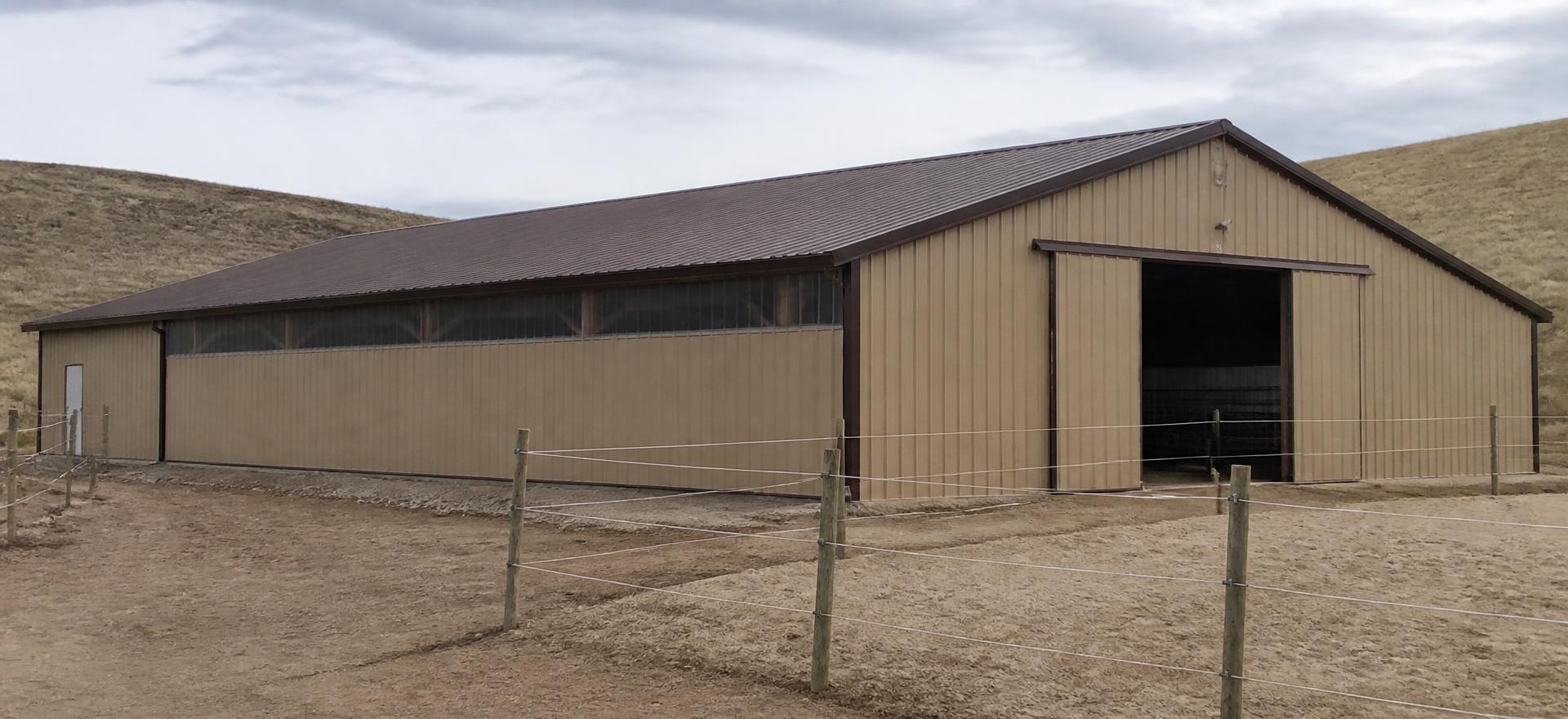 Concept photograph of a tan clear-span steel riding arena with a brown gable roof and a large open end door beside a sand paddock; example configuration, not a real project.