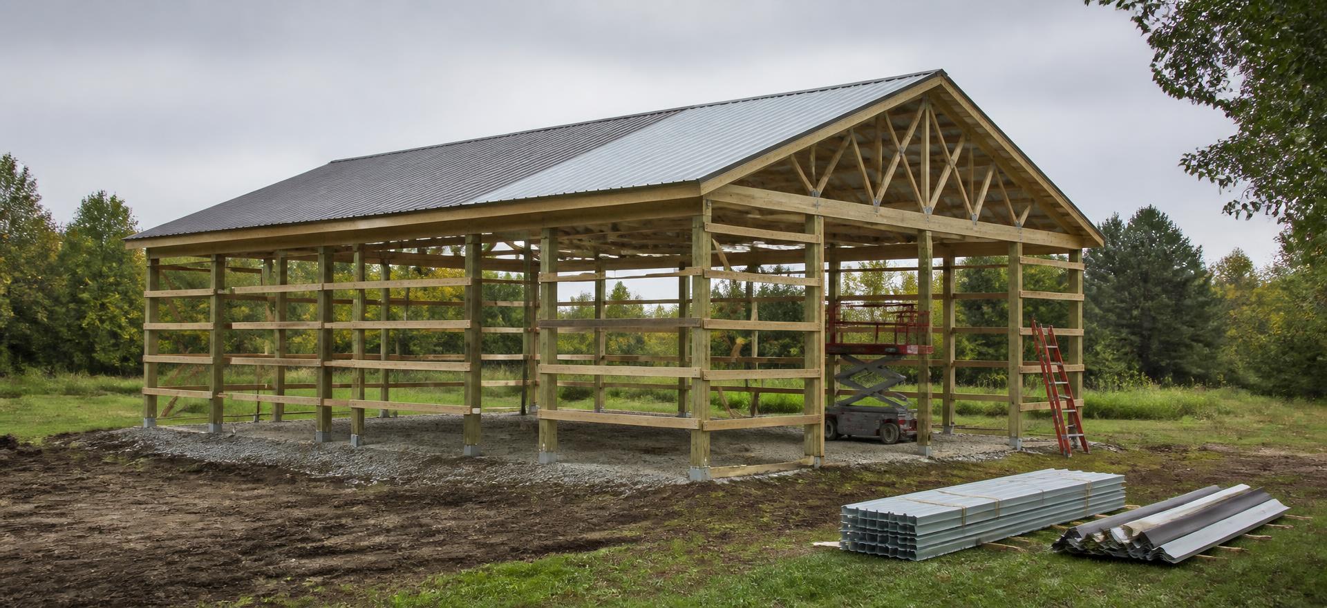 Concept photograph of a 36 by 48 foot post-frame building kit part-way through assembly, with treated columns, wood trusses, purlins and partly installed steel roof panels beside stacks of trim; illustrative configuration, not a real project.