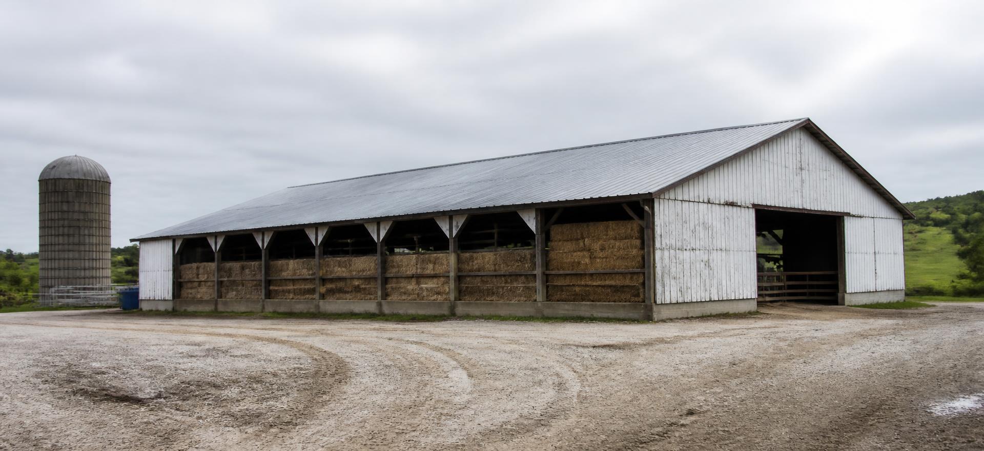 Concept photograph of a weathered white post-frame barn clad in steel with open sidewall bays stacked with hay bales beside a gravel yard; example configuration, not a real project.