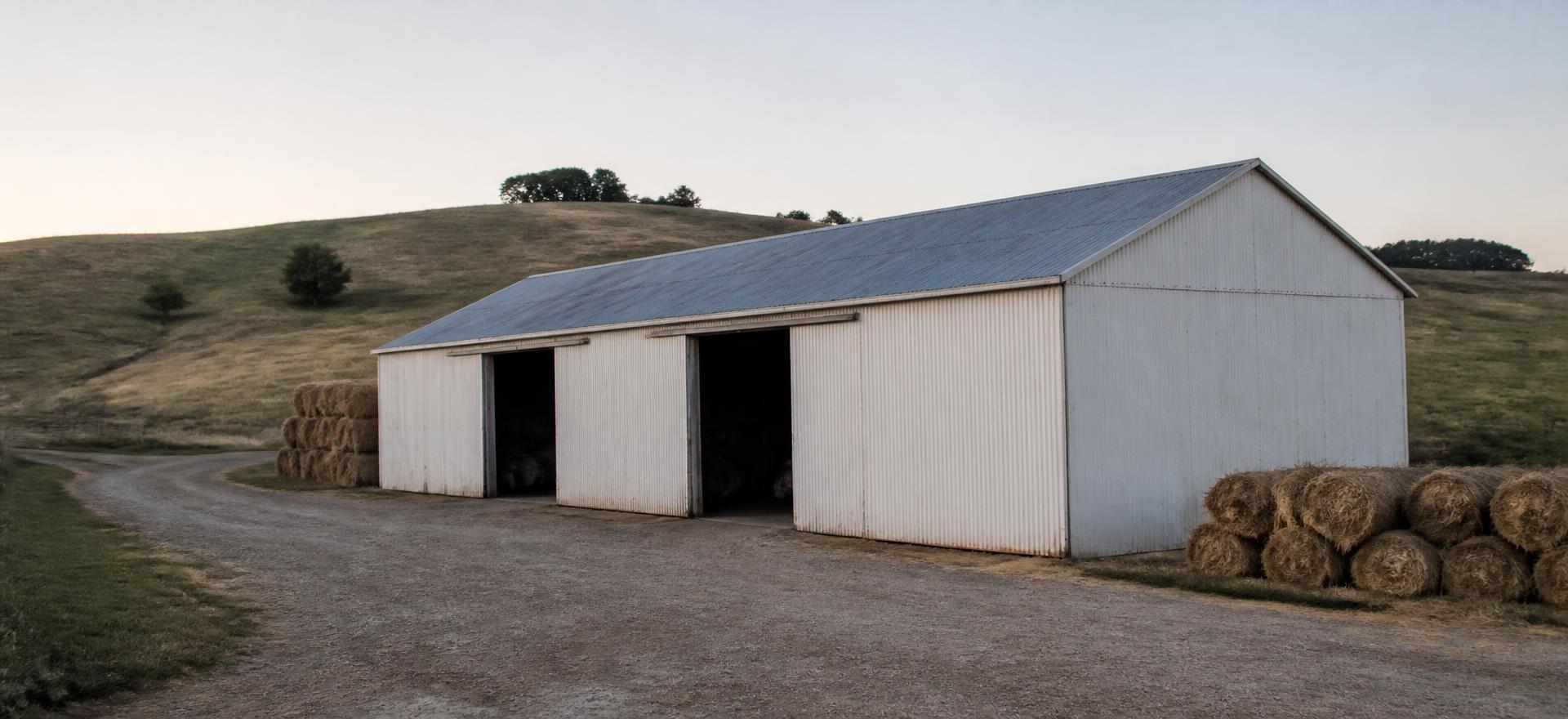 Concept photograph of a white steel-clad machine shed with two large sliding doors and round hay bales stacked outside; example configuration, not a real project.