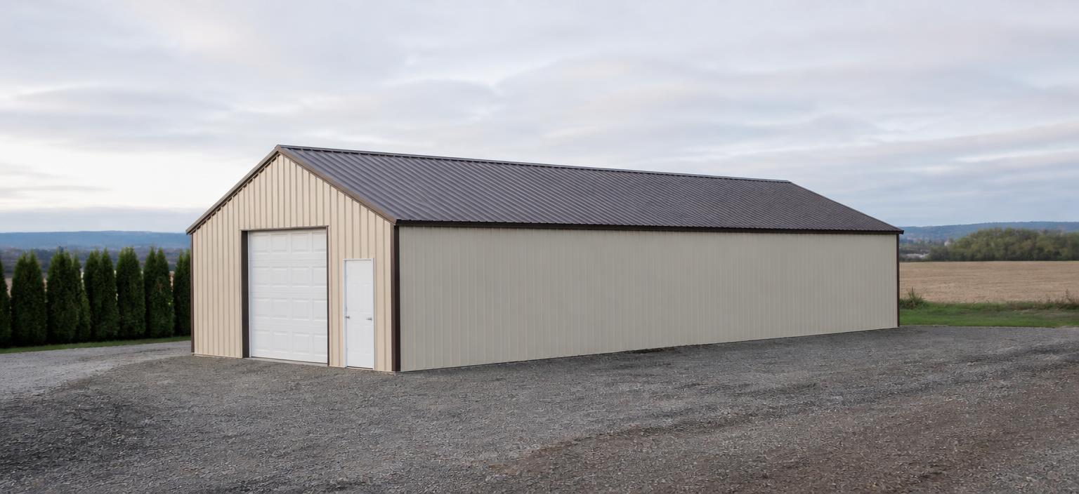 Illustrative steel building configuration showing a long beige steel storage building with one overhead door, one personnel door and a brown ribbed gable roof on a gravel yard beside farmland under soft cloud.