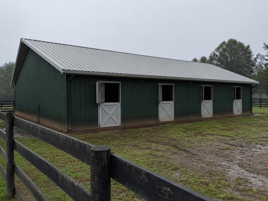 Concept photograph of a dark green steel-clad stable with four dutch stall doors facing a fenced paddock; example configuration, not a real project.