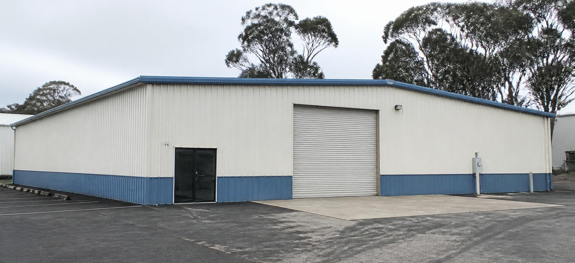 Concept photograph of an off-white steel warehouse with a blue base wainscot, one roll-up door and a glazed entry on an asphalt yard; example configuration, not a real project.