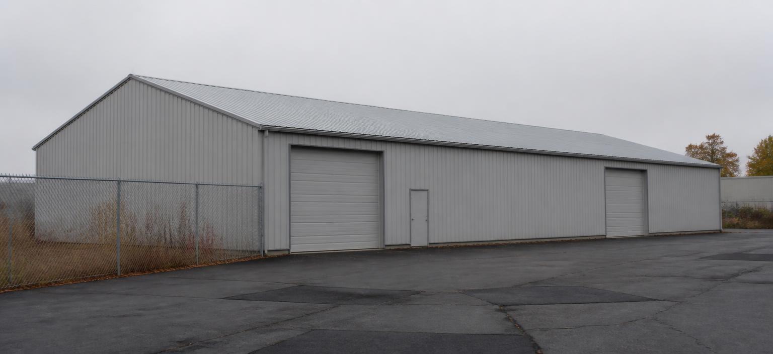 Illustrative steel building configuration showing a wide light grey warehouse with two overhead doors, one personnel door, a galvalume low-slope gable roof, chain-link fencing and patched asphalt under flat overcast light.