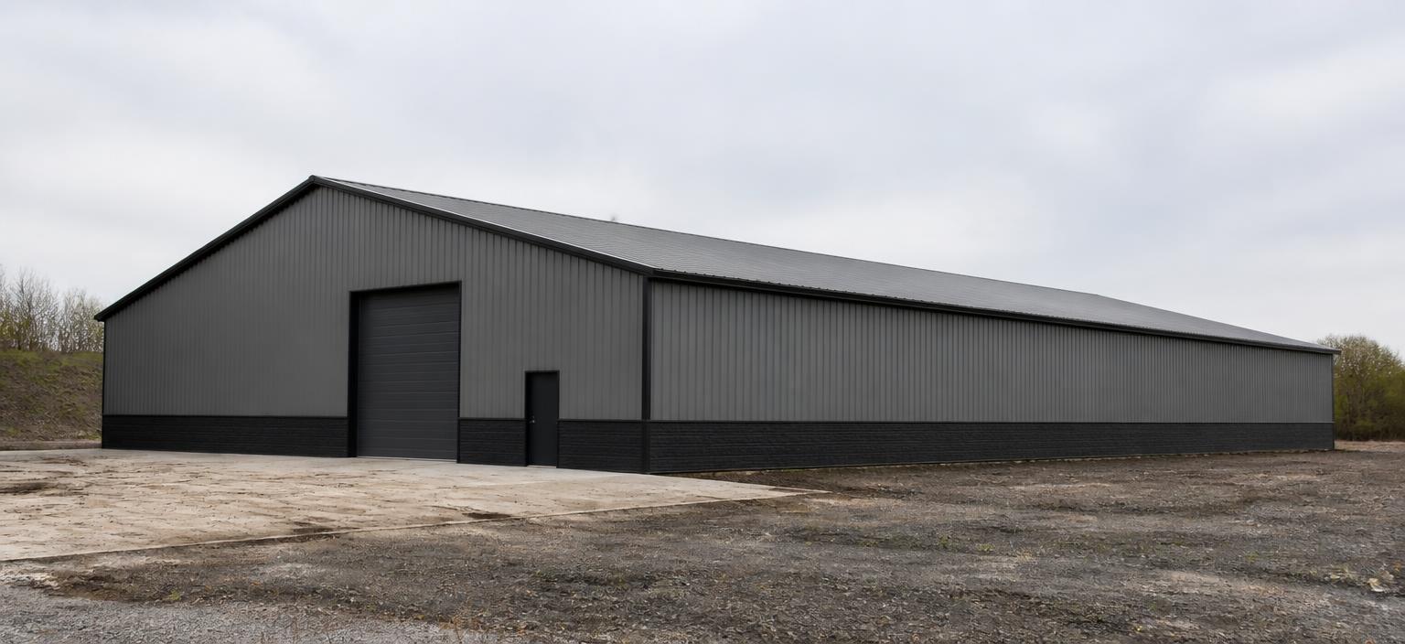 Illustrative steel building configuration showing a wide grey industrial steel building with a dark base course, one tall overhead door, one personnel door and a grey low-slope gable roof on a mixed gravel and concrete site under overcast sky.