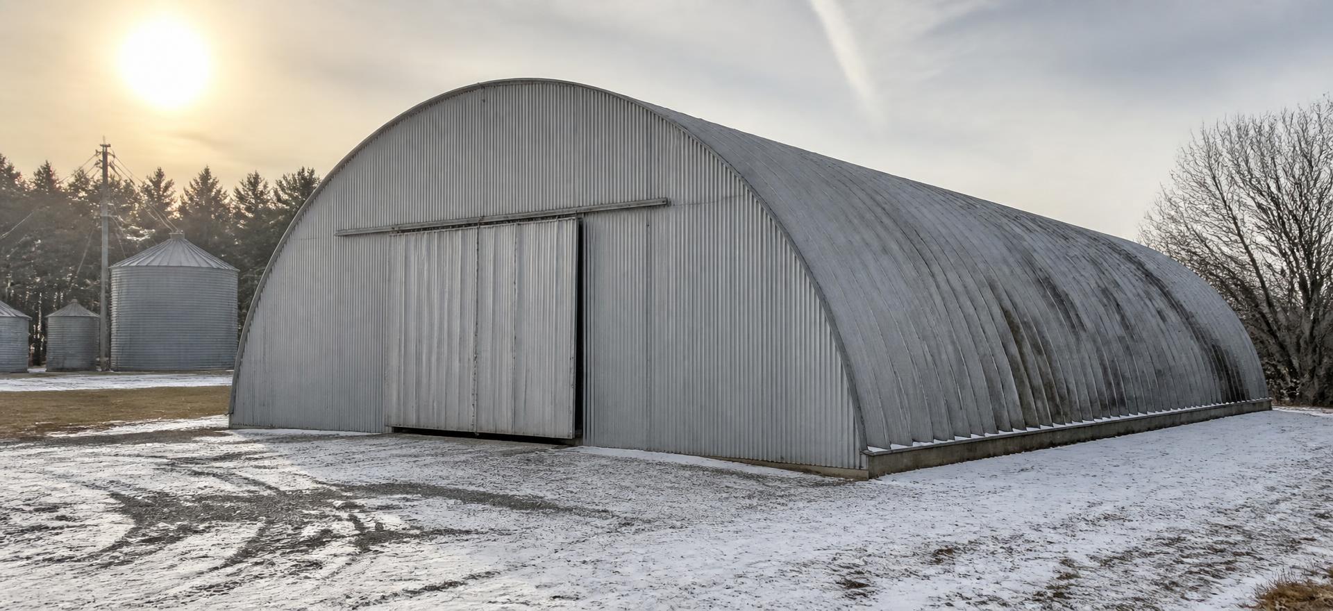 Concept photograph of a galvanized arch steel farm building with a flat sheeted end wall and a wide sliding door on a snow-dusted yard beside grain bins; example configuration, not a real project.