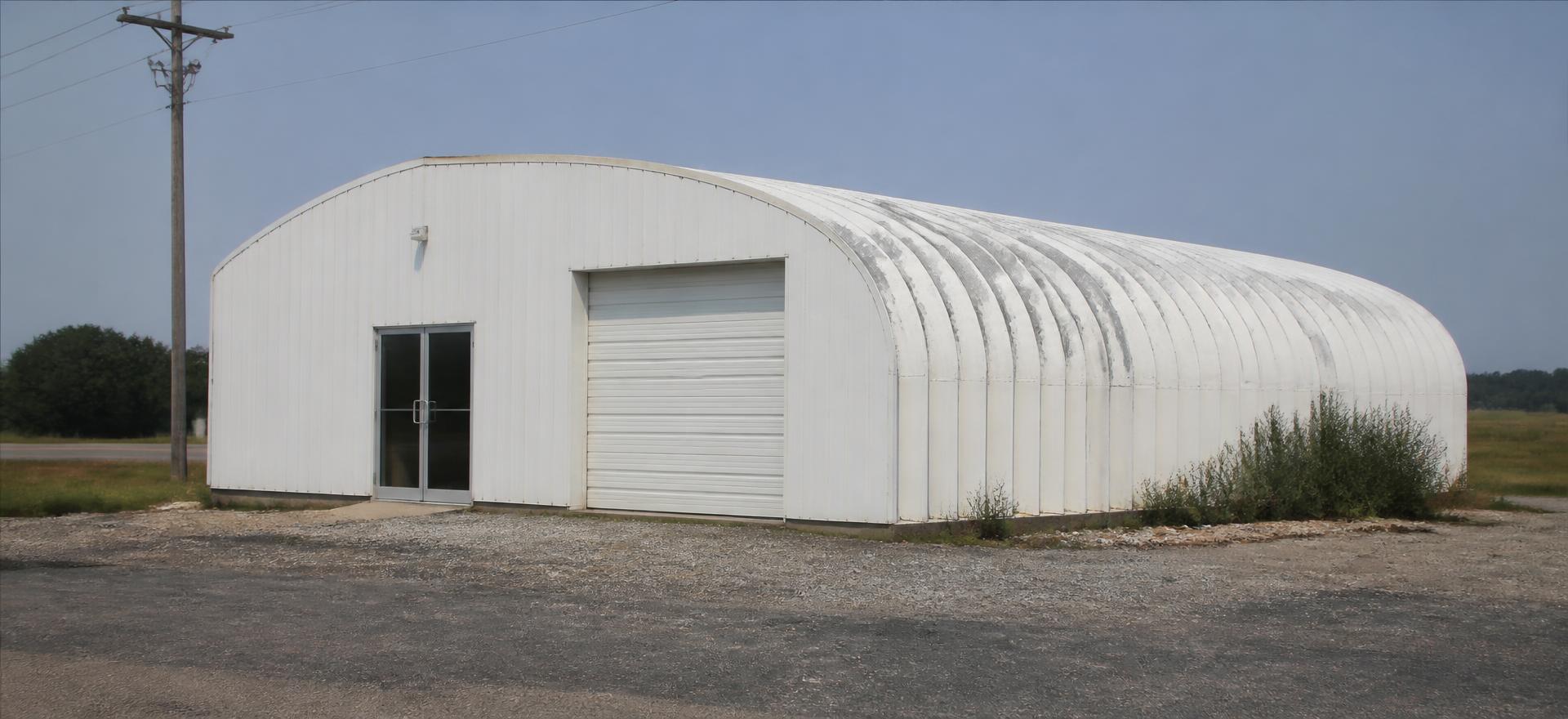Concept photograph of a white arch steel building with a squared front wall, a glass entry door and one roll-up service bay beside a gravel parking strip; example configuration, not a real project.