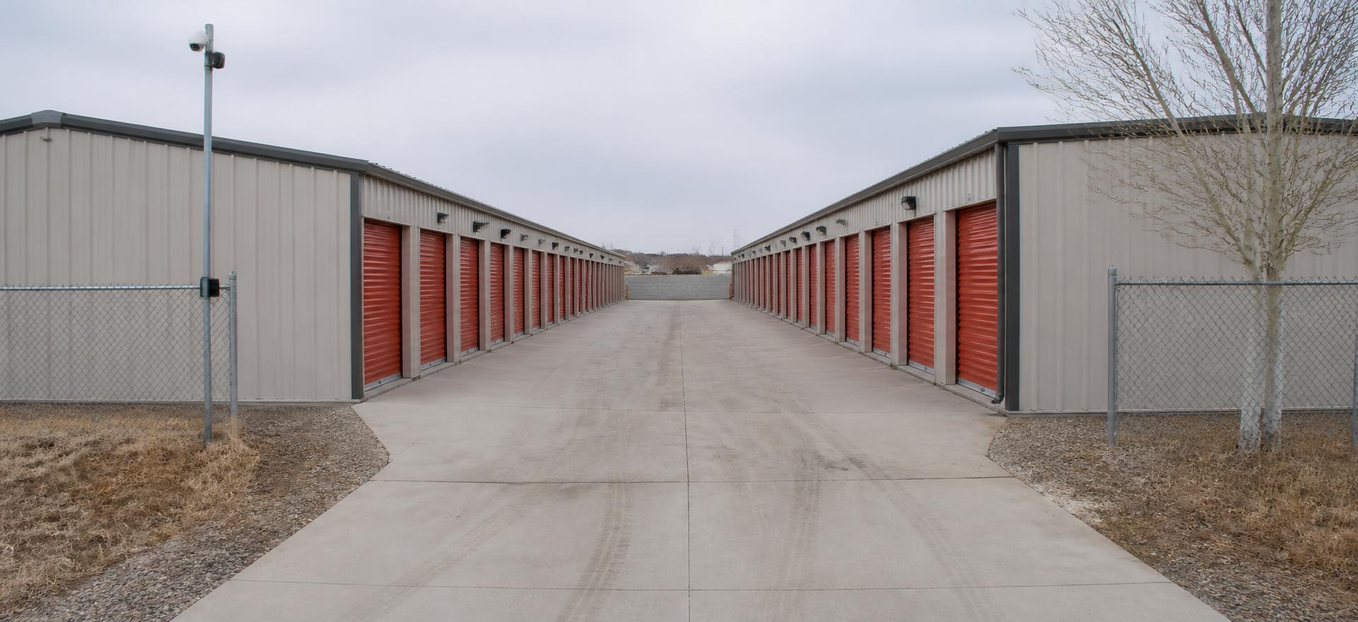 Concept photograph of two facing rows of single-story steel self-storage buildings with orange roll-up doors along a concrete drive aisle; example configuration, not a real project.