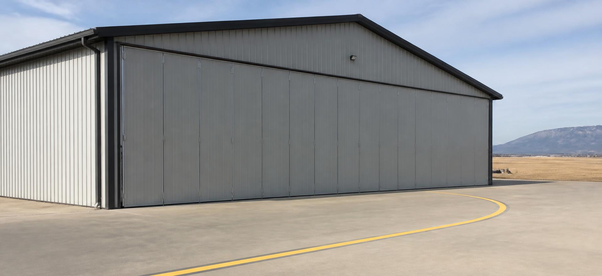 Concept photograph of a grey steel aircraft hangar with a full-width bi-fold door and a tall clear opening on a concrete apron with a painted taxi line; example configuration, not a real project.