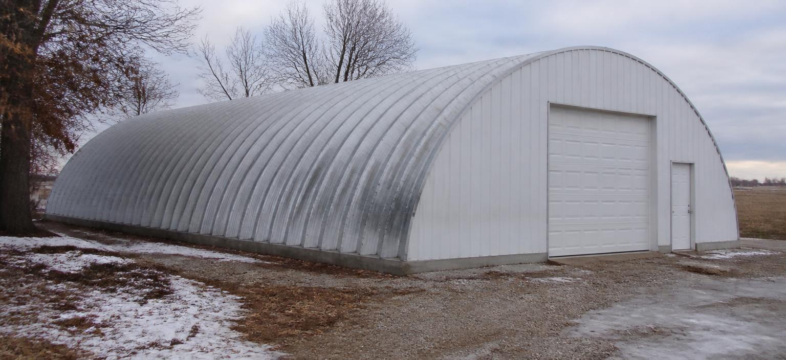 Steel building configuration showing a galvanized arch Quonset garage with a sheeted end wall, overhead door and walk door on a snow-patched gravel yard.