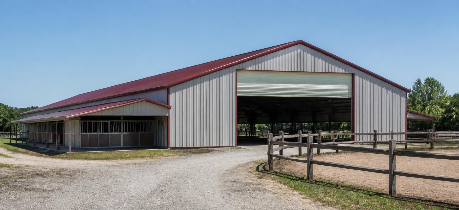 Concept photograph of a grey steel riding arena with a burgundy roof, a tall open gable bay and an adjoining stall wing beside a sand paddock; example configuration, not a real project.