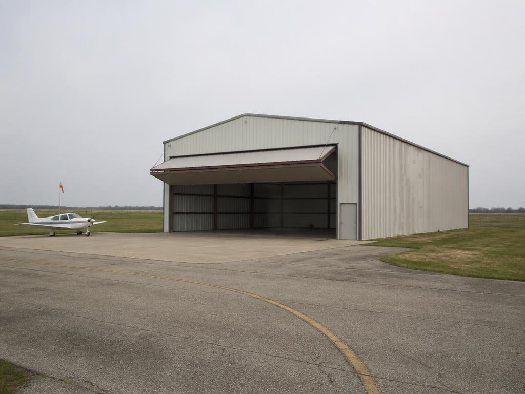 Concept photograph of an 80 by 100 foot clear-span steel aircraft hangar with a partly open bi-fold door on a concrete apron beside a taxiway; example configuration, not a real project.