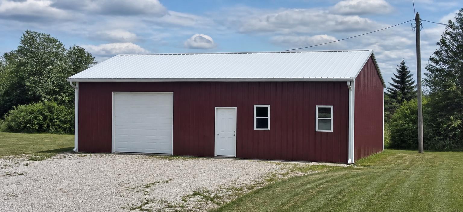 Illustrative steel building configuration showing a narrow burgundy steel workshop with white trim, one overhead door, one personnel door, two small windows and a white vertical-rib roof on a gravel pad beside mown grass.