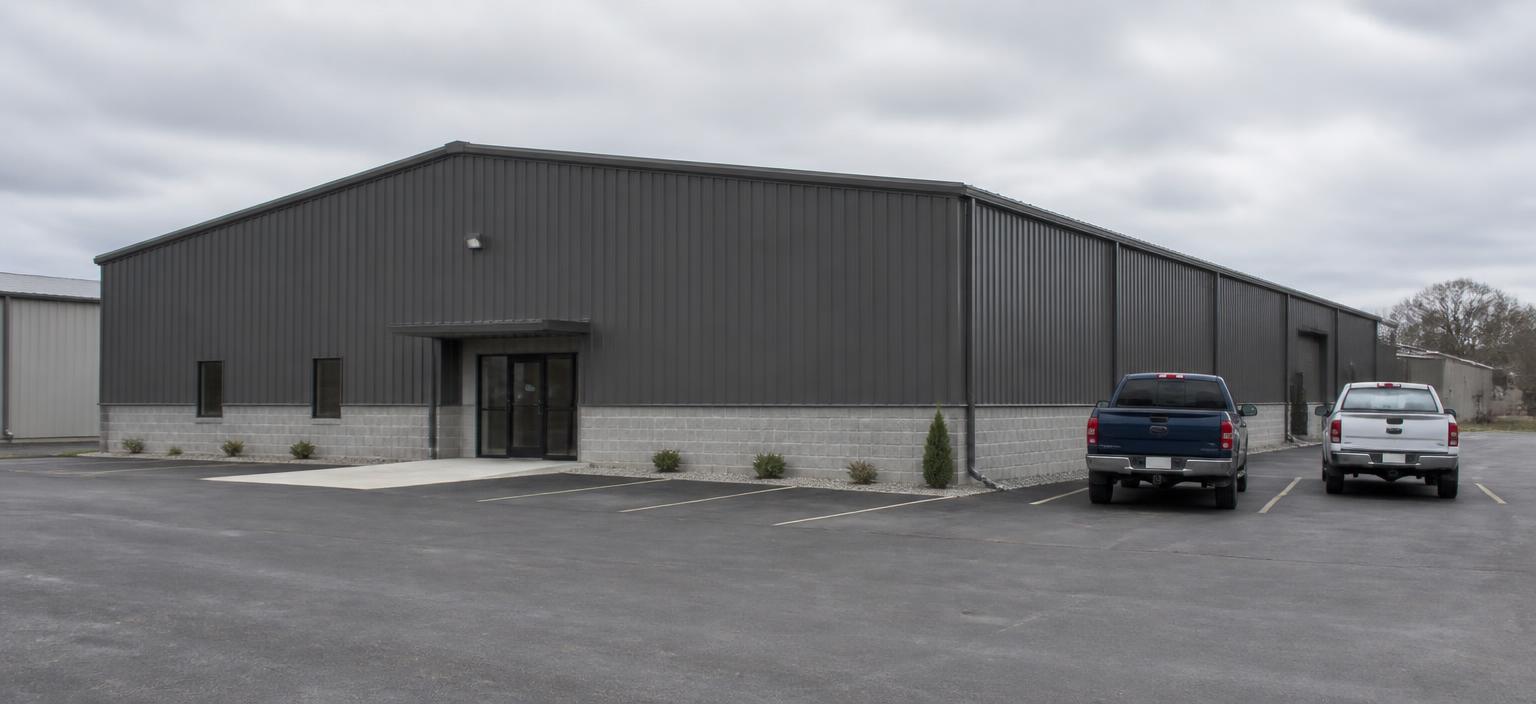Steel building configuration showing a charcoal rigid-frame commercial building with a masonry block wainscot and a small glazed entry beside a striped asphalt lot.
