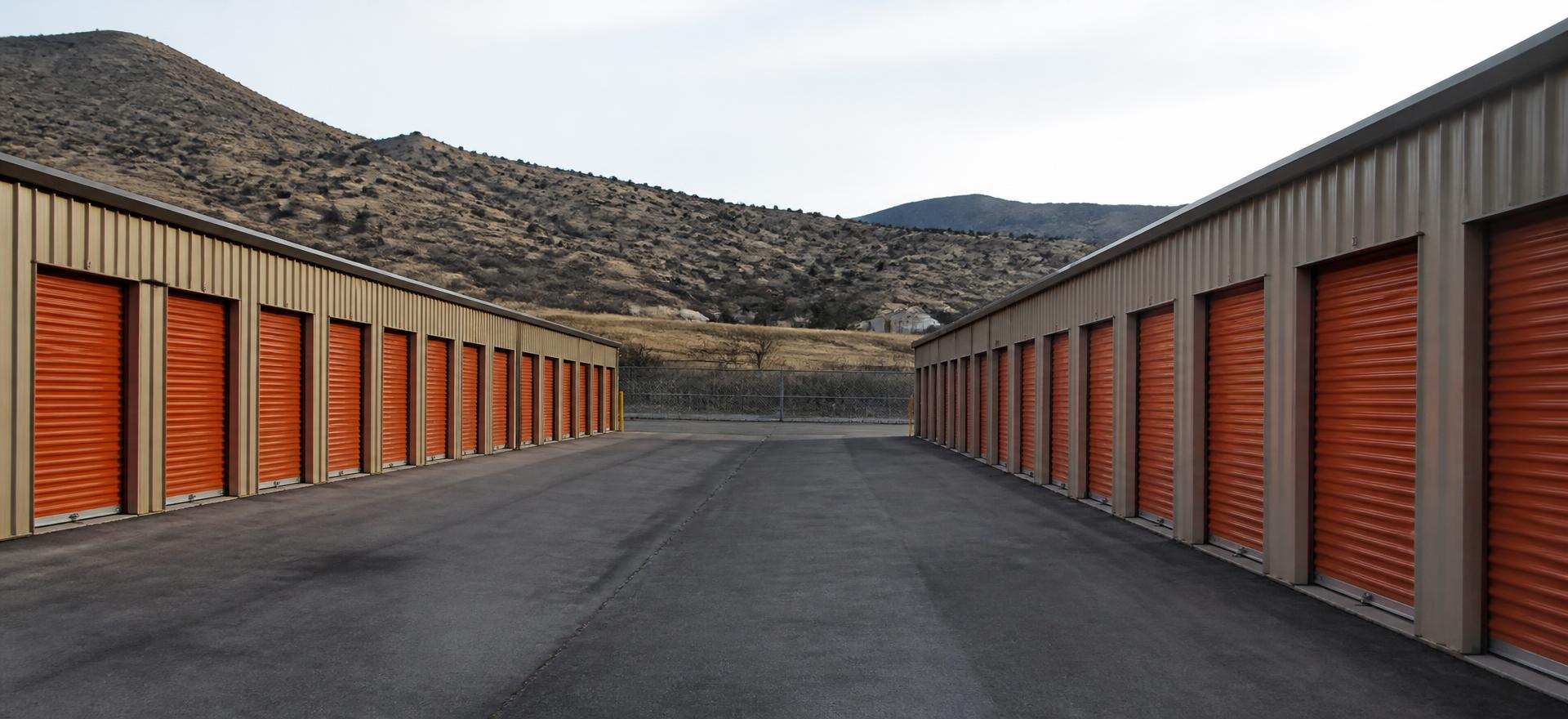 Concept photograph of two rows of tan steel self-storage buildings with orange roll-up unit doors either side of an asphalt drive aisle; example configuration, not a real project.