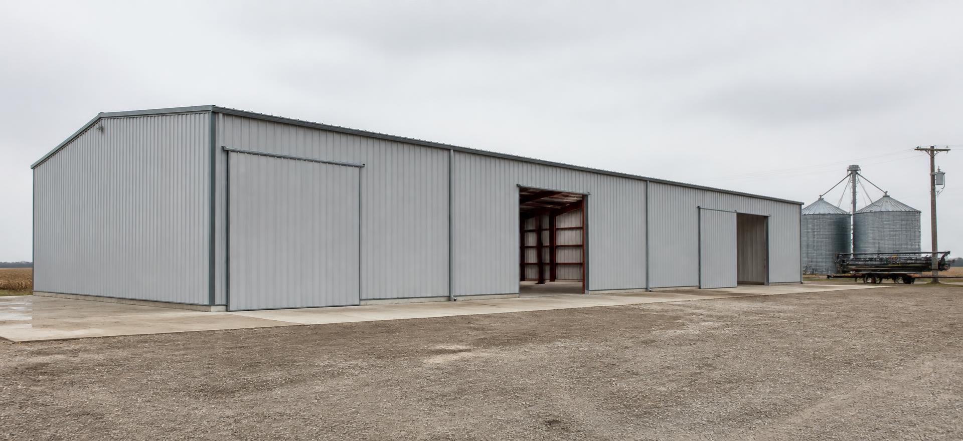 Concept photograph of a 60 by 150 foot gray steel agricultural equipment building with large sliding doors, one open showing primed interior framing, beside grain bins; example configuration, not a real project.